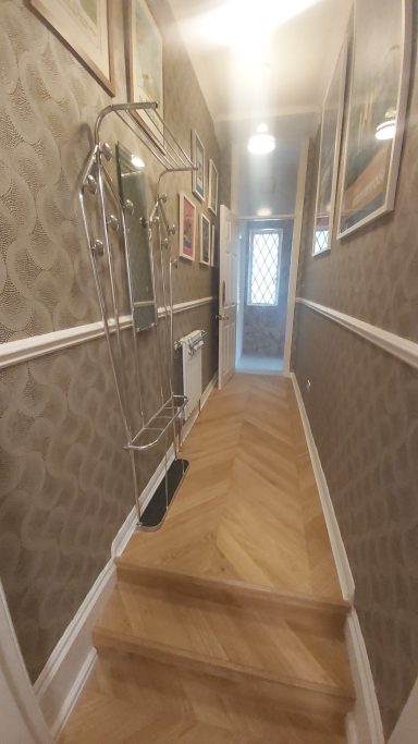 Hallway with grey patterned wallpaper, oak flooring and pictures hanging on the walls 